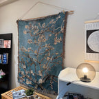 Woven wall art depicting floral branches hanging on a wall next to a shelf with books and a lamp.